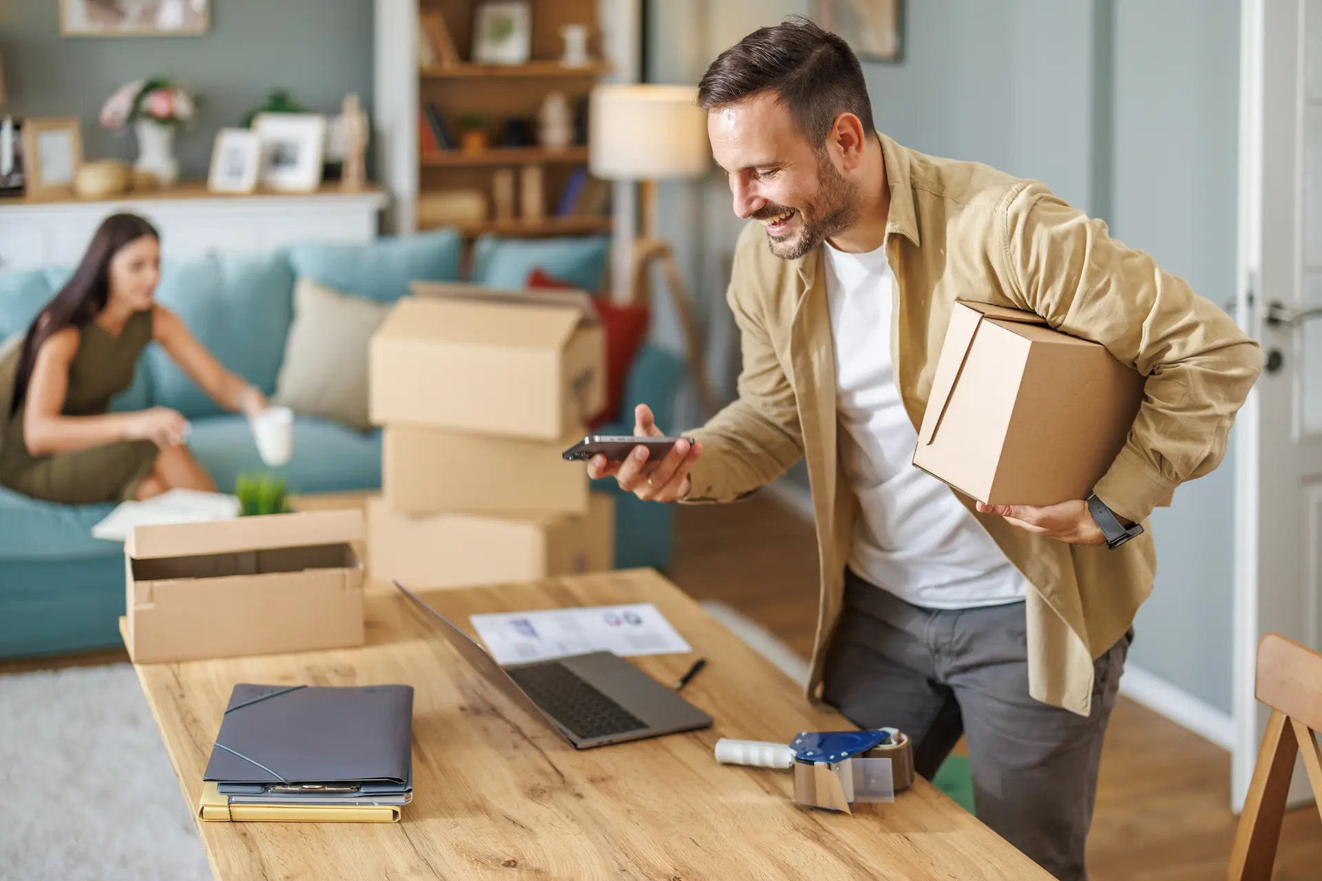 Smiling entrepreneur carrying a shipping box while checking his phone in his workspace.